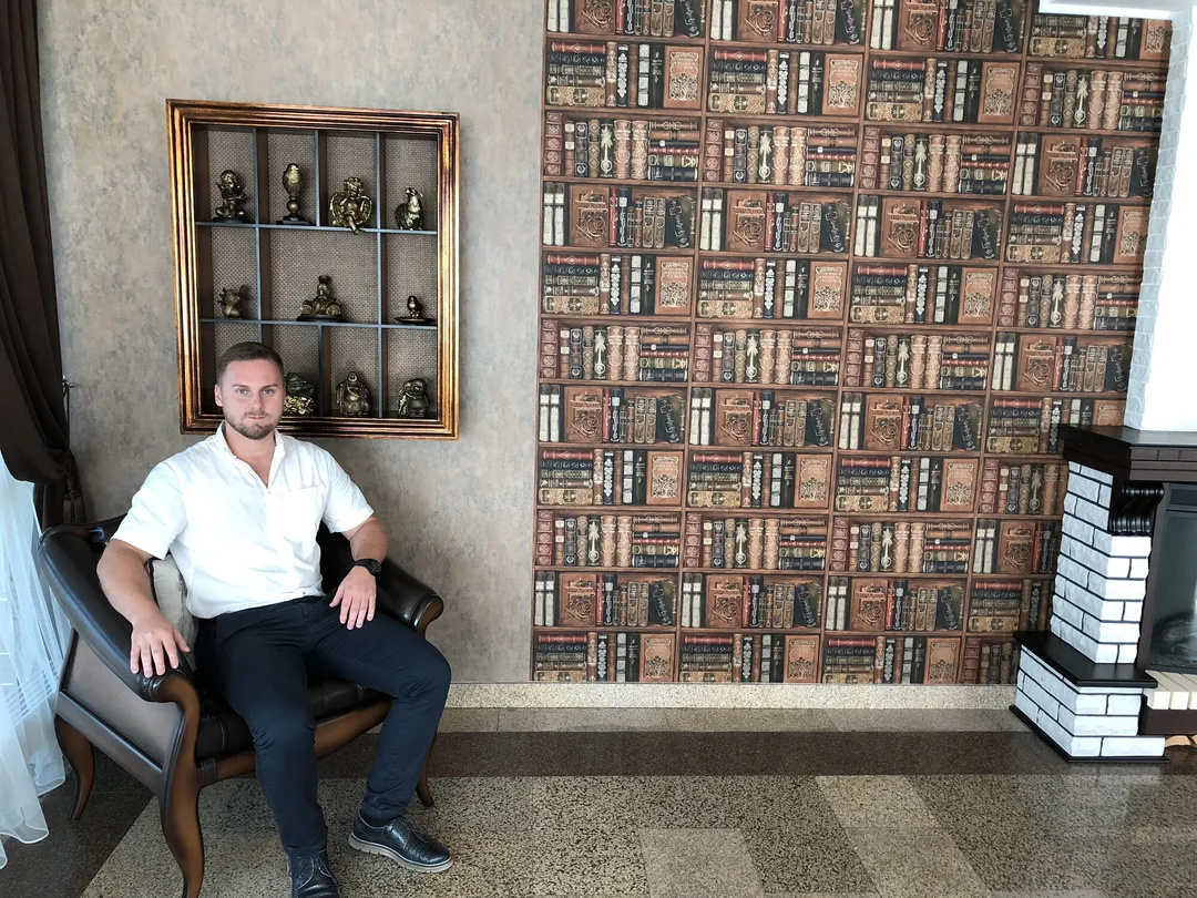 Дмитрий Котов в кресле в интерьере с книжными полками
